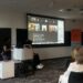 Image of the front of the room: Brooklyn and Shane next to the Zoom screen with Tom Shakespeare presenting online.