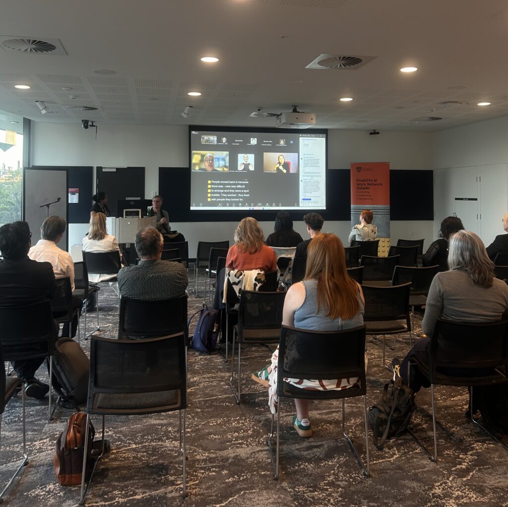 Image taken from the back of room: audience members seated facing the Zoom screen with Tom Shakespeare presenting online.
